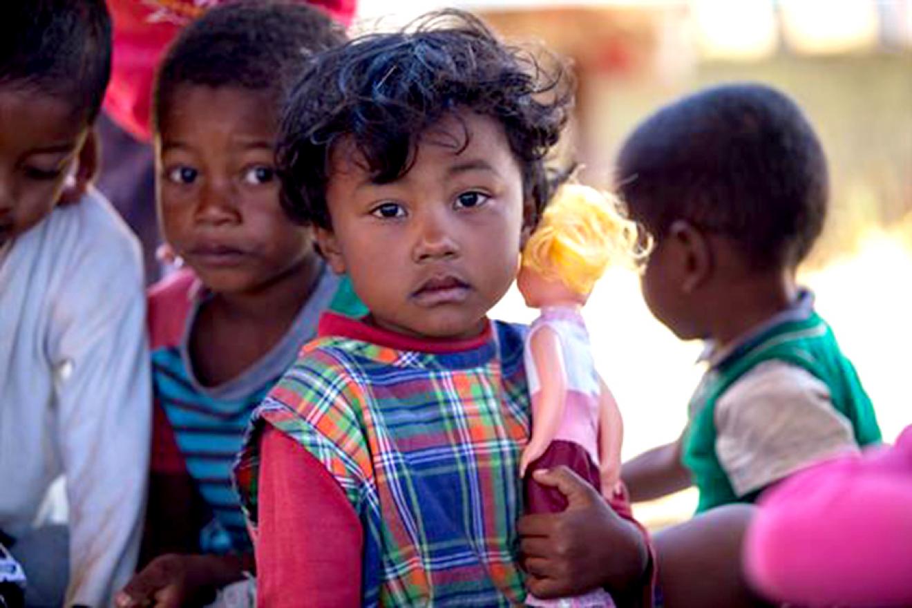 A child holding a doll among other children faces the camera.